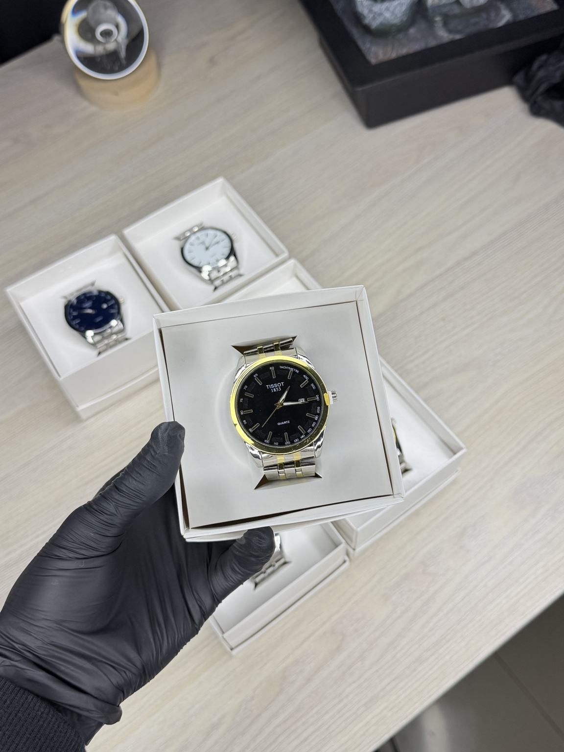 Mano con guante sosteniendo una caja blanca con un reloj de acero y detalles dorados, con más relojes en cajas al fondo.